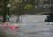 Severe urban flooding: cars partially submerged in brown floodwater on a street, with a bridge and buildings in the background.