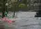Severe urban flooding: cars partially submerged in brown floodwater on a street, with a bridge and buildings in the background.