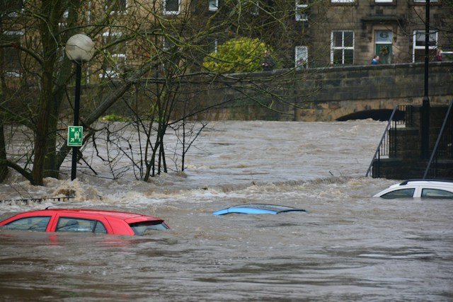 Severe urban flooding: cars partially submerged in brown floodwater on a street, with a bridge and buildings in the background.