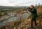 Hunter in camouflage aims a scoped rifle on a rocky autumn hillside as a gray dog runs ahead.