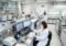 Scientist in a white lab coat and mask pipettes liquids at a workbench in a modern chemistry lab with computers and bottles nearby.