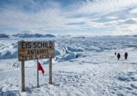 Wooden sign in Antarctic ice reads 'EIS SCHILD ANTARKIS 79°15'S, 41°20'E' as a group of explorers in red jackets approach in a vast icy landscape.