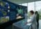 Scientists in a modern command center study a wall of climate data panels and interact with a touchscreen console; wind turbines are visible outside the windows.