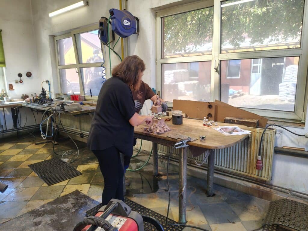 Woodworker carving small statues at a cluttered workbench in a workshop, with hoses and tools visible in the background.