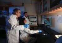 Three researchers in lab coats, two standing and one seated, analyze a computer monitor showing a blue?and?red graph.