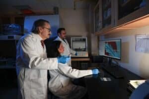 Three researchers in lab coats, two standing and one seated, analyze a computer monitor showing a blue?and?red graph.