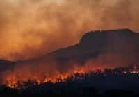 Wildfire burns along a forested hillside at dusk, with orange smoke-filled sky and glowing embers along the treeline.