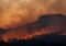 Wildfire burns along a forested hillside at dusk, with orange smoke-filled sky and glowing embers along the treeline.