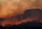 Wildfire burns along a forested hillside at dusk, with orange smoke-filled sky and glowing embers along the treeline.