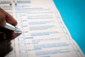 Close-up of a hand using a pen to mark a vote on a German ballot paper with party names like Grüne and FDP on a blue surface above it