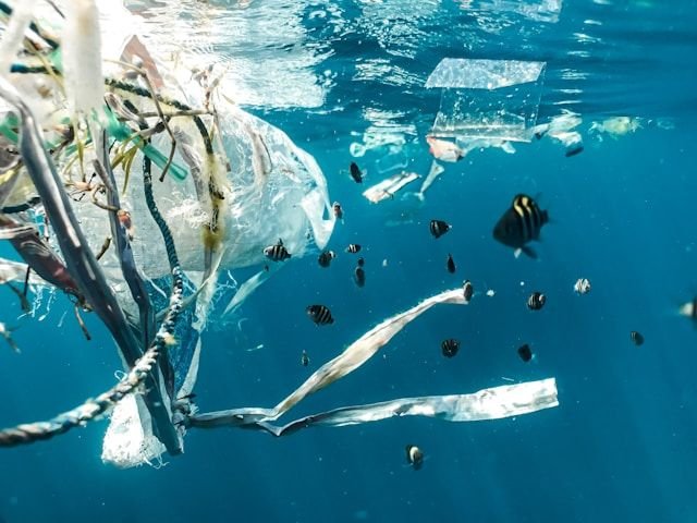 Underwater view of plastic bags and trash drifting among small tropical fish in clear blue water, highlighting ocean pollution.