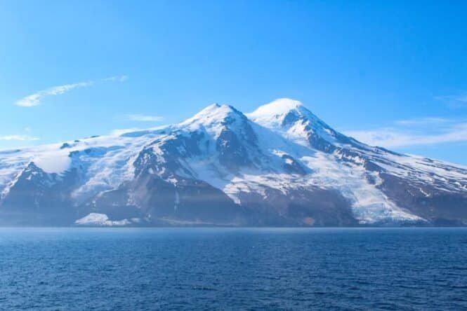 Snowy mountain range rising from a calm blue fjord under a clear sky