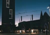 Evening city street with a tall dark tower showing a dot?matrix clock, tram wires above, and lit shopfronts along the sidewalk.