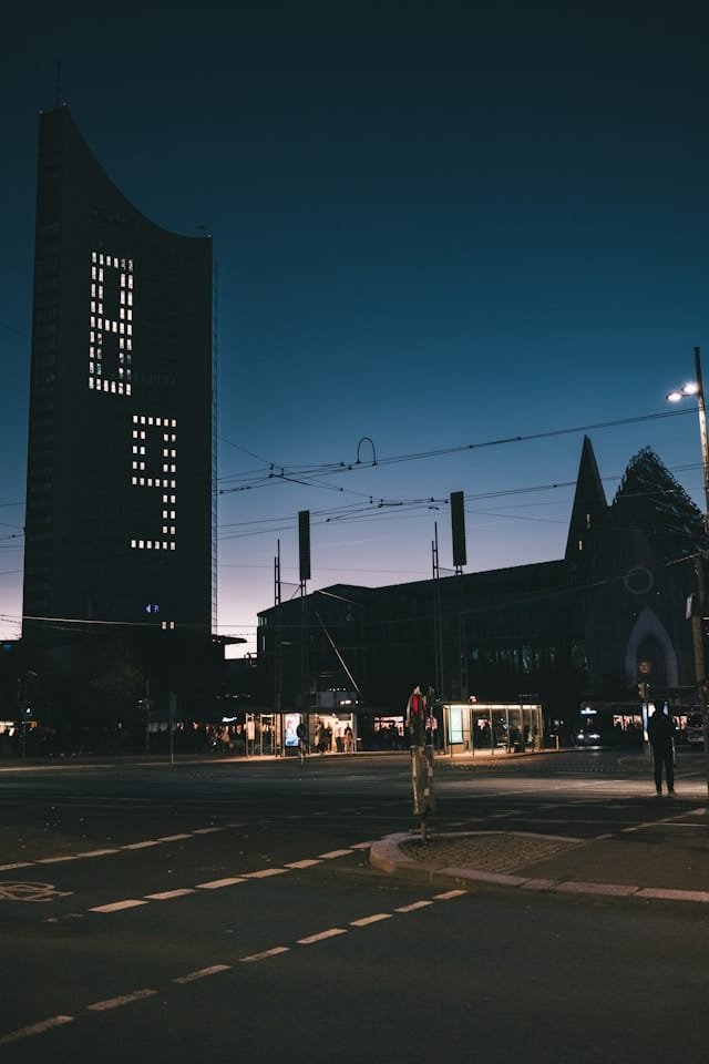 Evening city street with a tall dark tower showing a dot?matrix clock, tram wires above, and lit shopfronts along the sidewalk.