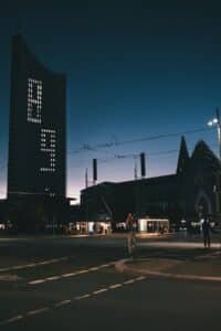 Dusk city street with a tall left-building displaying dotted lights, tram wires overhead, and people by a lit storefront.