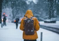 Person seen from behind wearing a bright orange winter coat and a dark backpack, walking through falling snow in a city street.