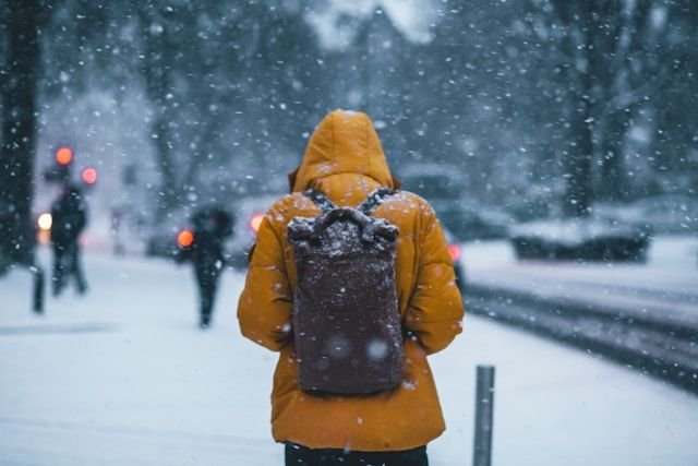 thom-holmes-9gbwzrbt0qu-unsplash6447765943648161456jpg | Pugnalom Person seen from behind wearing a bright orange winter coat and a dark backpack, walking through falling snow in a city street.