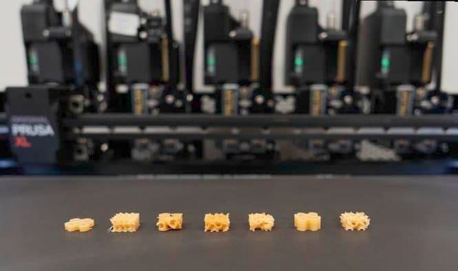 Row of seven small beige 3D-printed gear-shaped objects on a printer bed, with the machine blurred in the background.