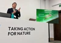 Speaker at a conference podium with a banner reading 'Taking Action for Nature' and a presentation screen beside him.