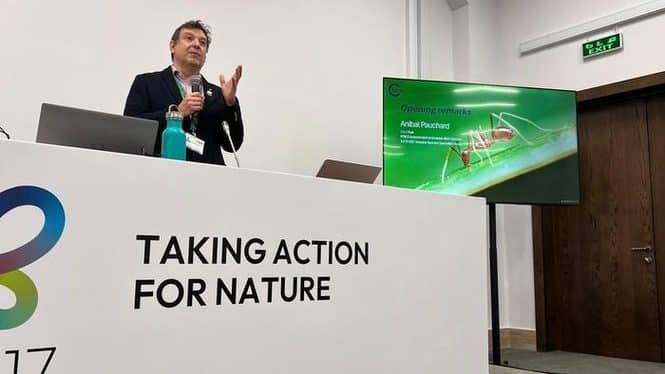 Speaker at a conference podium with a banner reading 'Taking Action for Nature' and a presentation screen beside him.