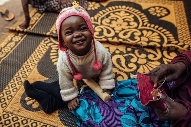 Young child wearing a pink knit hat smiles while sitting on a patterned rug as someone sews bright pink fabric nearby.