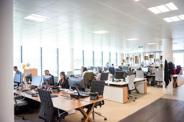 Open-plan office with employees working at desks and computers under bright ceiling lights.