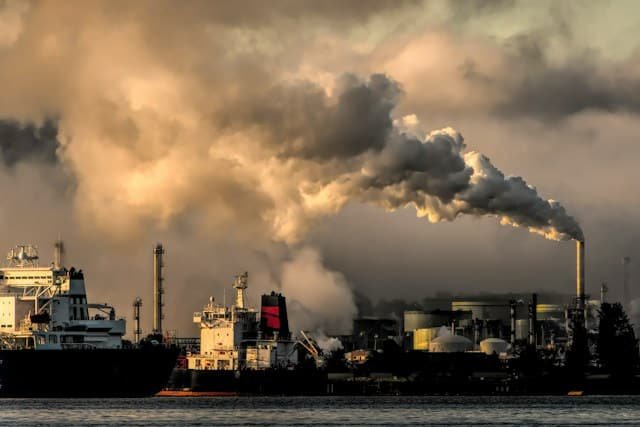 Industrial refinery or factory by the water releasing thick smoke from tall chimneys into a cloudy sky at dusk.