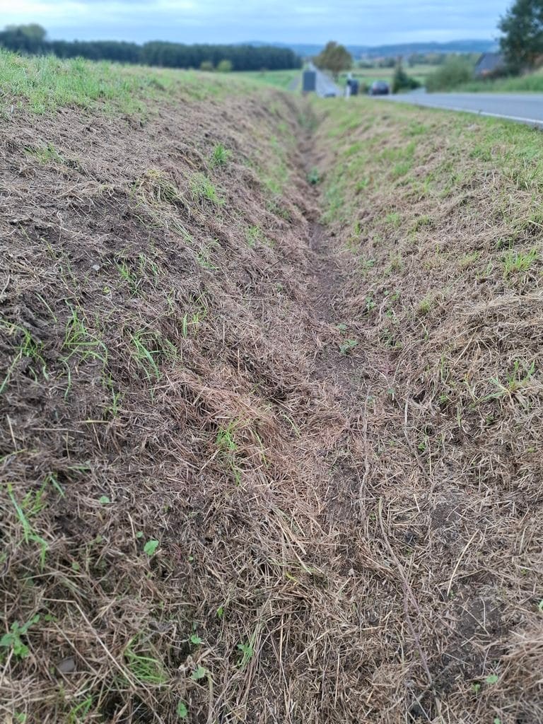 Deep dirt ditch along a rural roadside with brown dried grass and patches of green, extending into the distance under a cloudy sky.