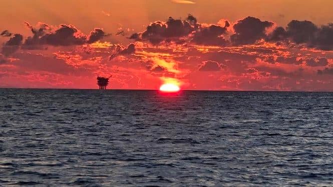 Sunset over the ocean with a small tree silhouette on the horizon and a fiery orange sky.