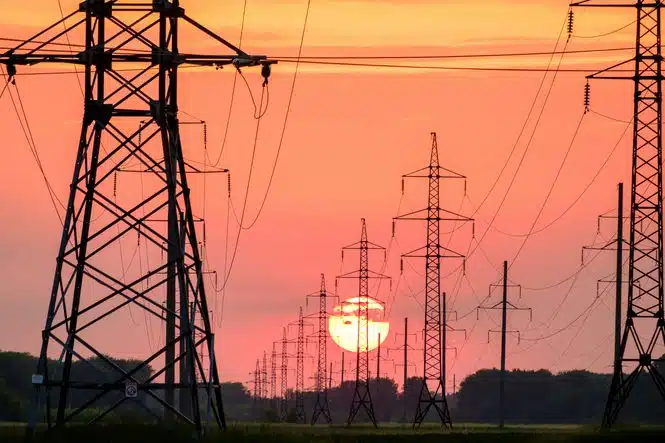Silhouetted transmission towers and power lines against an orange sunset sky.